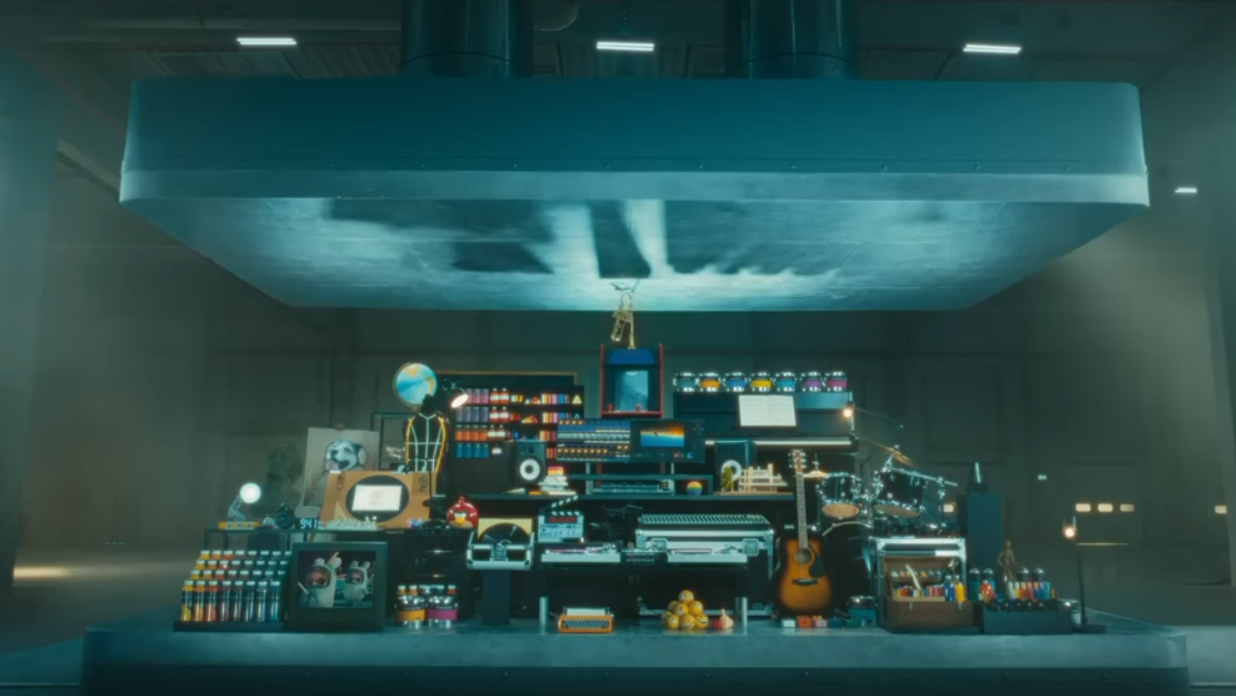 A still of the aforementioned Apple commercial. It's a room with a platform, and a roughly pyramid shaped assortment of various artsy things, many of which are musical instruments. There are guitars, drum kits, a globe, electric keyboards, a globe (for some reason?), cans of paint atop a piano, etc. Above all this is an ENORMOUS press, connected by two large pistons to some unseen contraption in the ceiling, coming down to crush everything